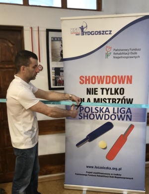 Na zdjęciu Prezes Zarządu Łukasz Skąpski, ubrany w klubową koszulkę, przecinający wstęgę na tle roll-upu projektu Showdown Nie Tylko Dla Mistrzów – Polska Liga Showdown.